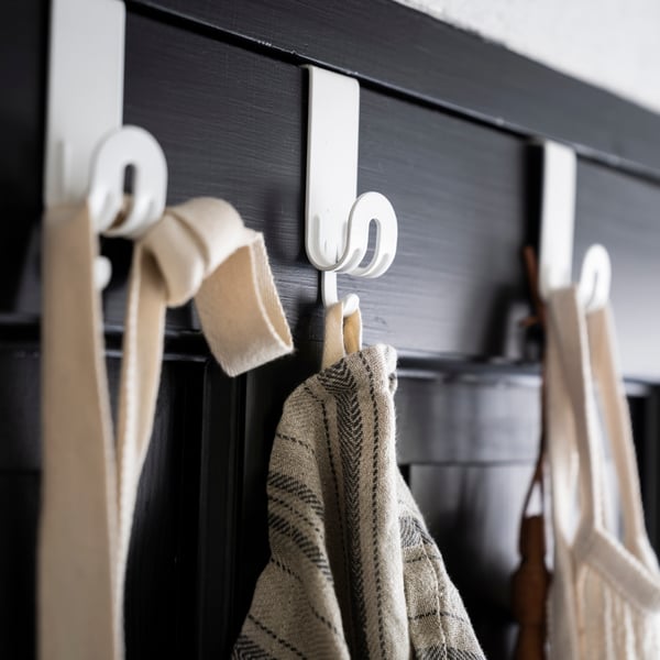 Three white SEKINER hooks for doors with various items such as a bag and an apron hanging from them.