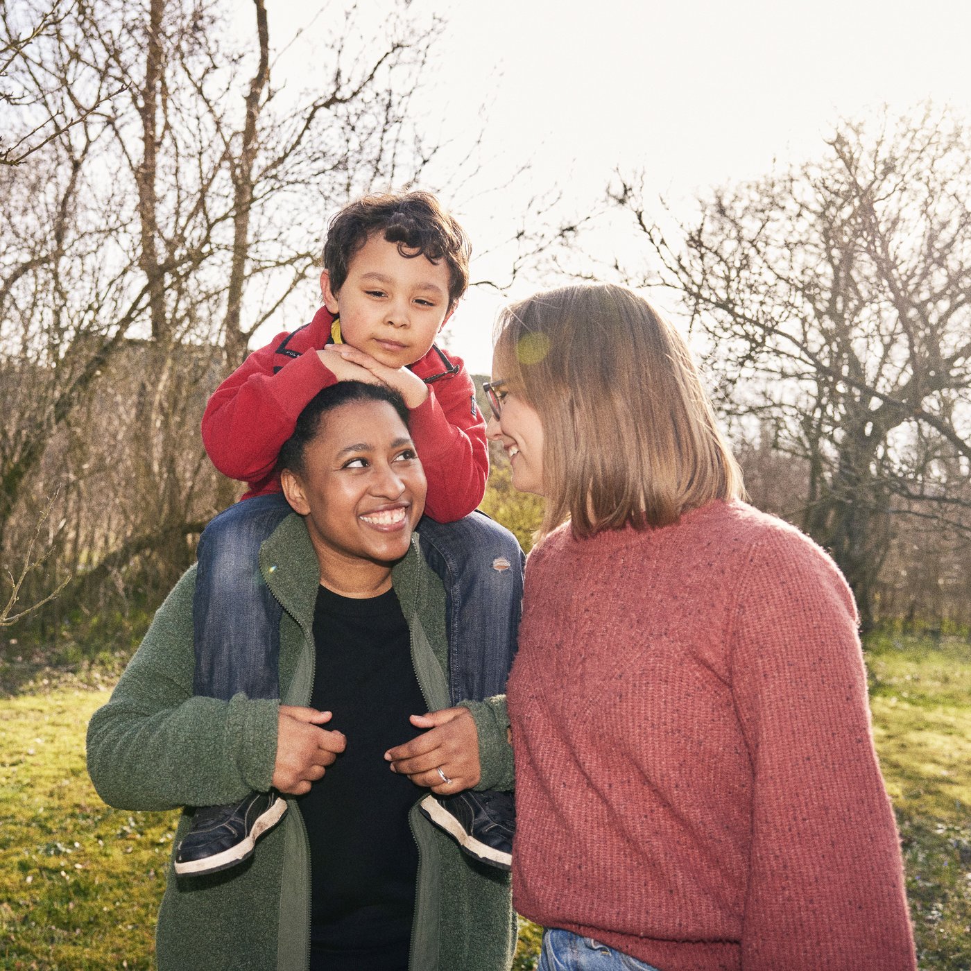 Alle and her wife standing outside in their garden, their son is sitting on her shoulders.