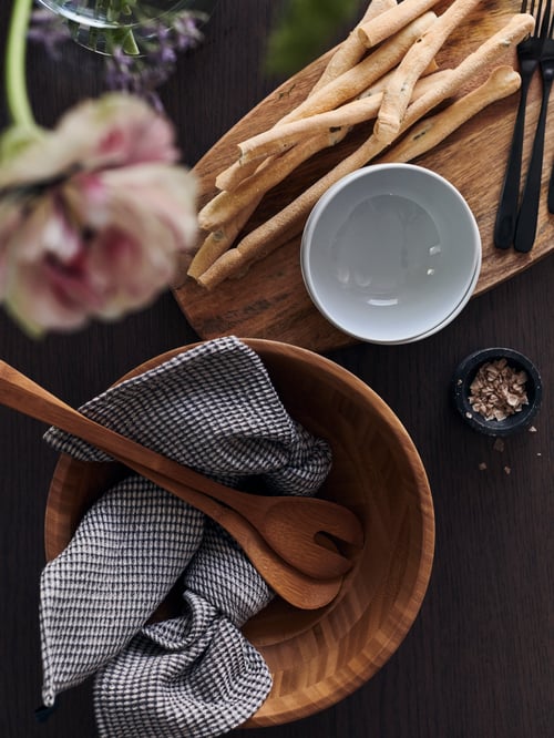 A GRÖNSAKER serving bowl with serving spoons, beside a chopping board with breadsticks, forks and a small bowl on it.