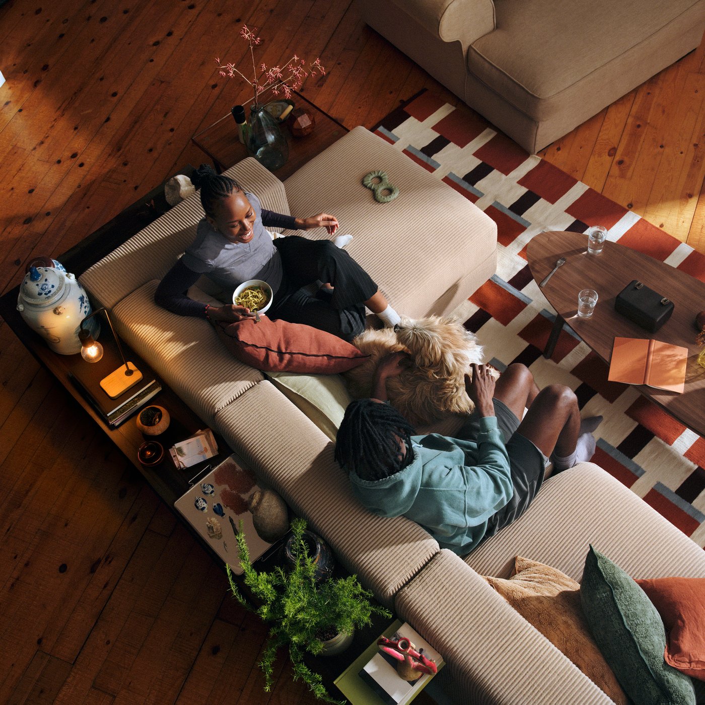 Two people are talking and sitting on a beige sofa with a dog, a colourful rug underneath.
