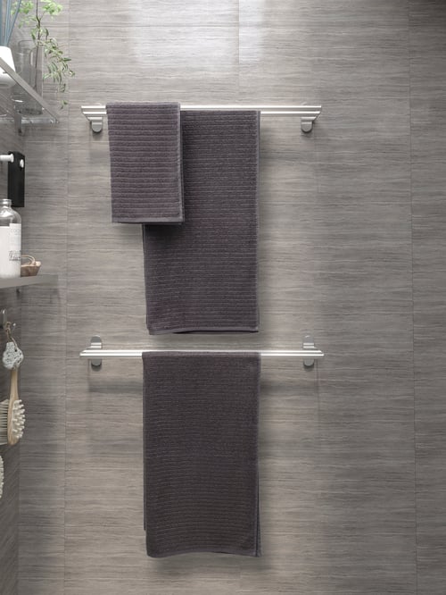 Salle de bains avec carreaux texturés, porte-serviettes, serviettes de toilette et serviettes de bain, tablette en verre, crochets, vase transparent, articles de toilette et brosse de douche.