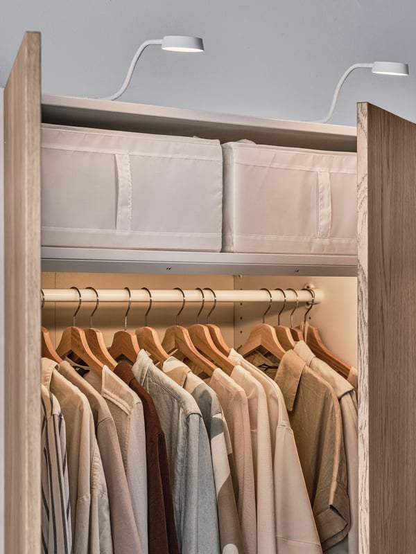Two white  SKUBB storage cases on the top shelf of a PAX/TONSTAD wardrobe with a hanging rail holding various clothes below.