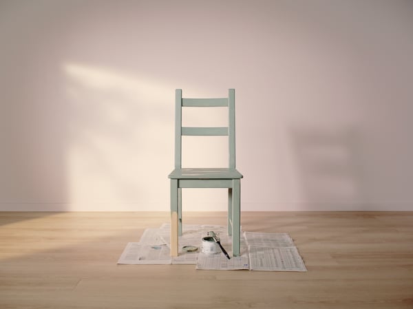 Une chaise IVAR peinte en vert clair posée sur de vieux journaux dans une pièce vide au parquet en bois clair et aux murs rose clair.