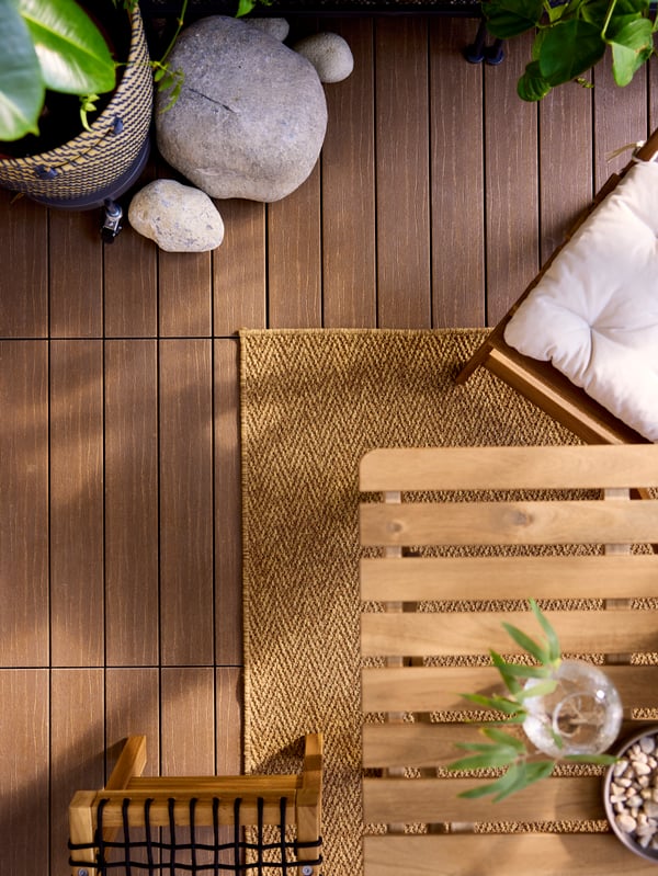 Wood-like brown UTLÄNGAN outdoor floor decking is seen from above under decorative stones, a woven rug and acacia furniture.
