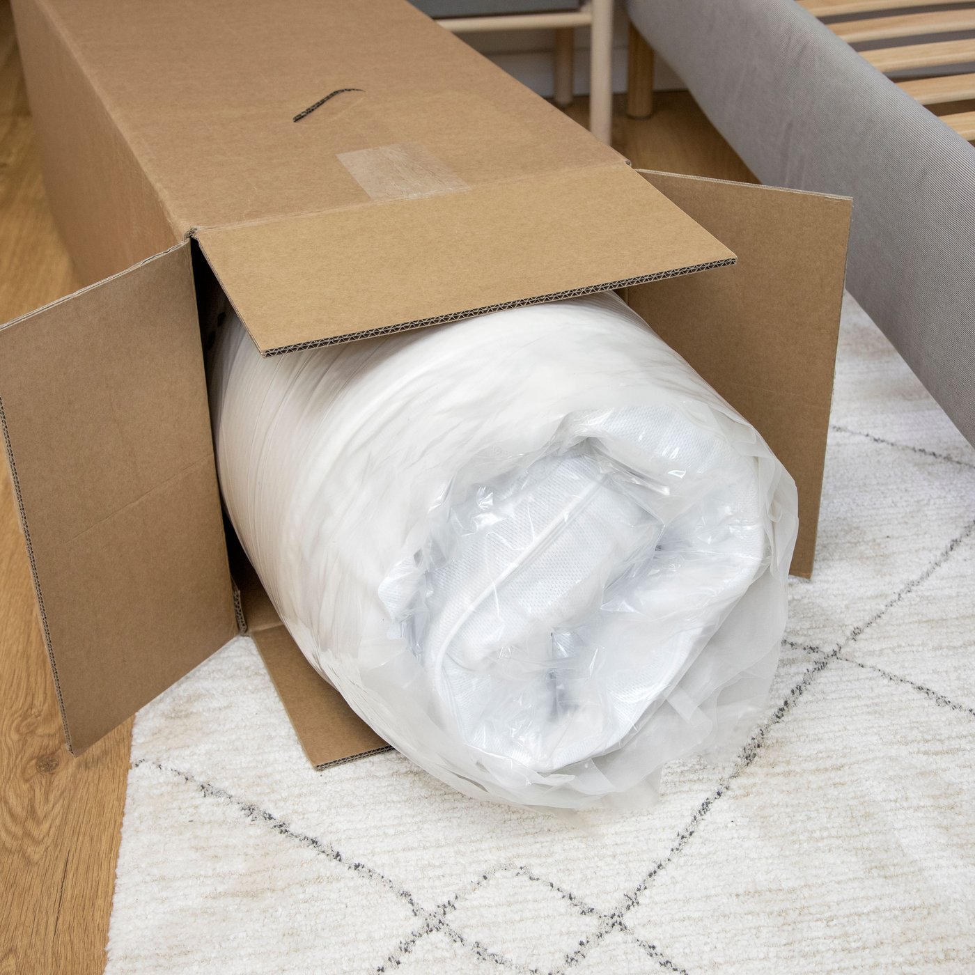 Matrace srolovaná ve fólii, částečně vytáhnutá z kartónové krabice na bílém koberci.