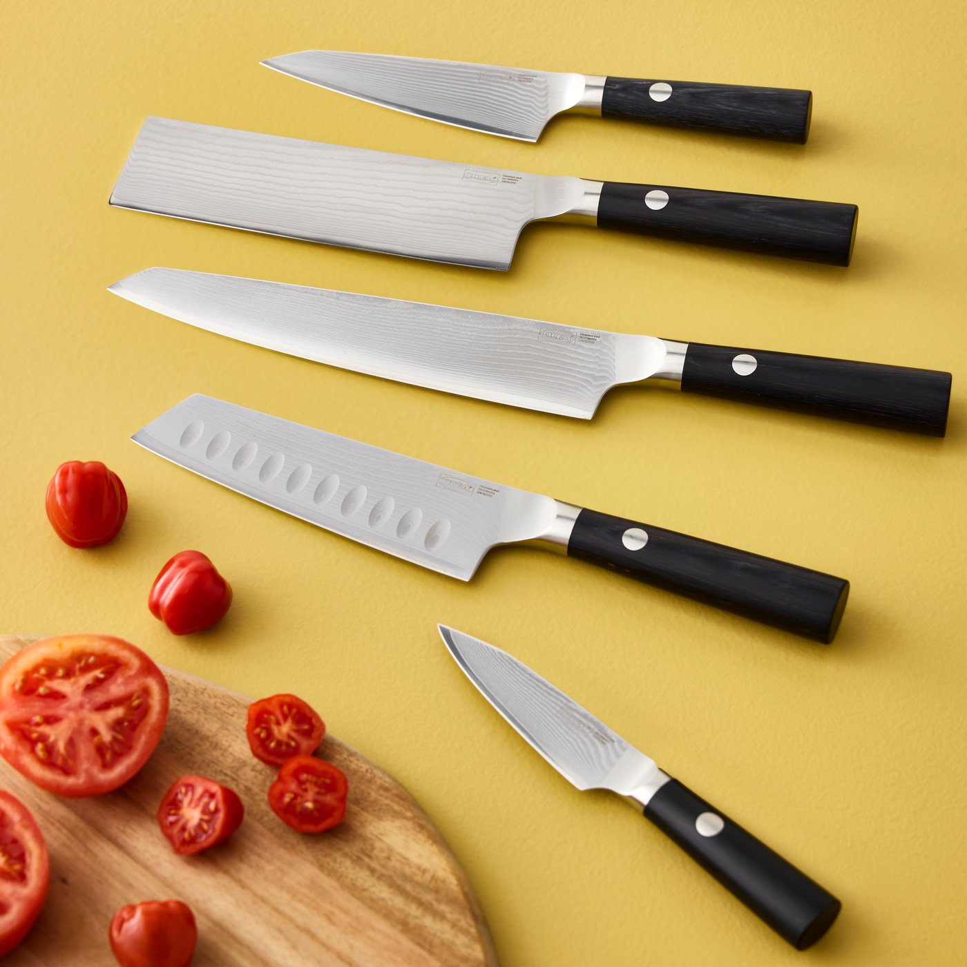 A close-up of five black FJÄLLRÖDING knives made of damascus steel on a yellow background with diced tomatoes.