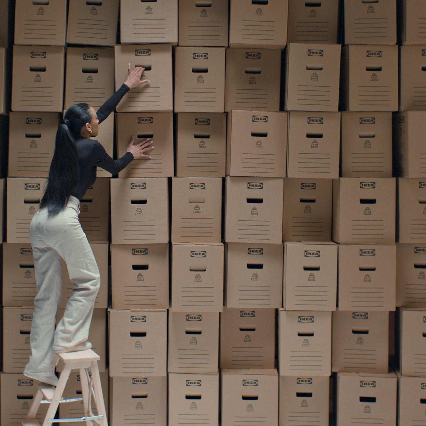 A woman stands on a step ladder on front of a wall of IKEA moving boxes