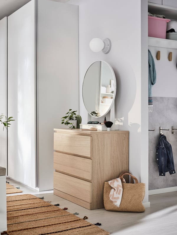 En MALM byrå i ljust trä, rund spegel, växter står i en hall. Till vänster om byrån står en vit garderob med skjutdörrar, och till höger ett öppet förvaringsutrymme med krokar, hyllor och kläder. På golvet ligger en vävd matta med tofsar samt en stråväska med filt.