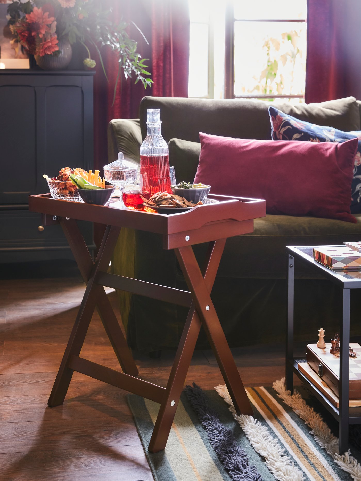 طاولة صينية حمراء داكنة بها وجبات خفيفة في سلطانيات وعصير في إبريق زجاجي، موضوعة على سجادة مخططة بجوار صوفا بلون أخضر زيتوني.