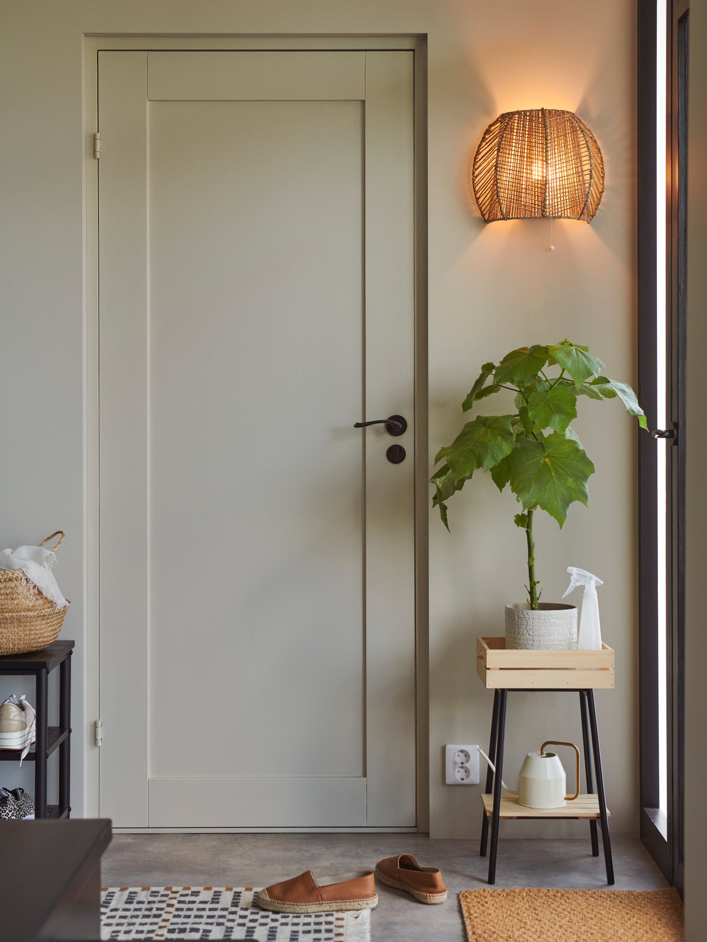 Vit hall där det står en piedestal med en grön växt intill en vit dörr. Ovanför piedestalen sitter en tänd vägglampa i naturmaterial.
