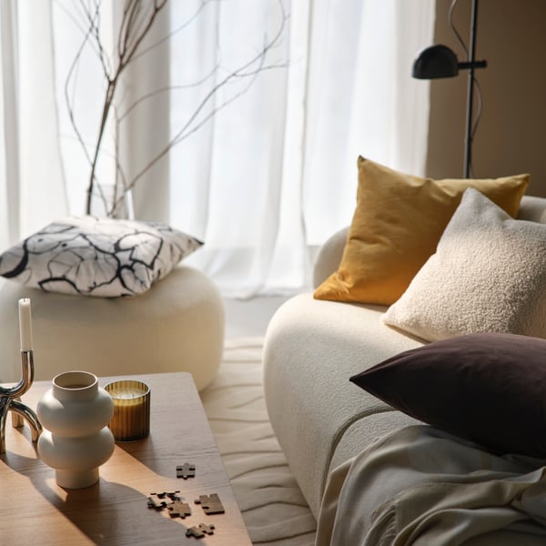 A light beige sofa with SANELA cushion covers and a PERSIKOTRÄD throw, by a coffee table with TOFSTÄRNA scented candle.