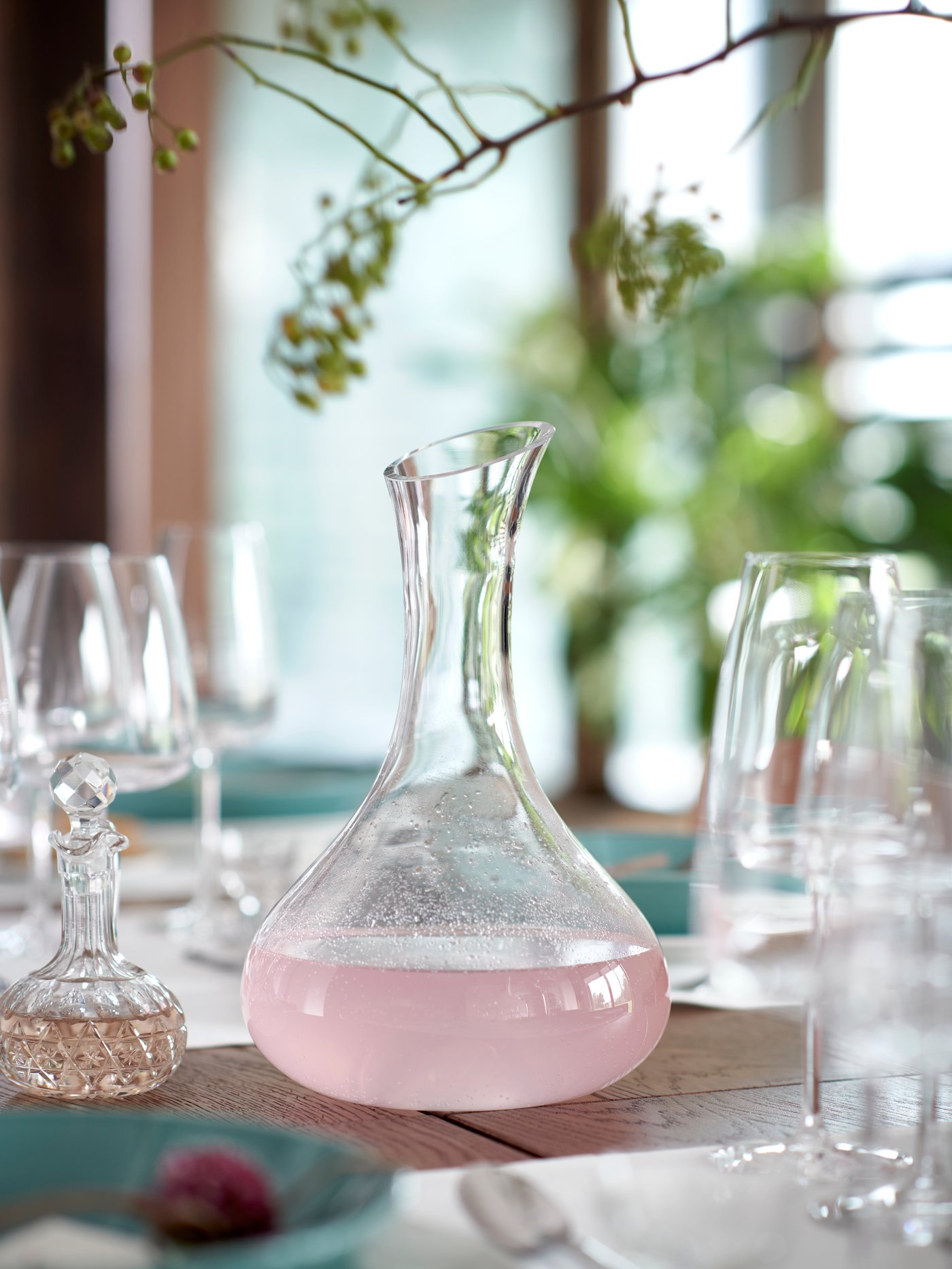 A carafe sits on a table filled with clear glasses. Above it hangs a decorative branch. 