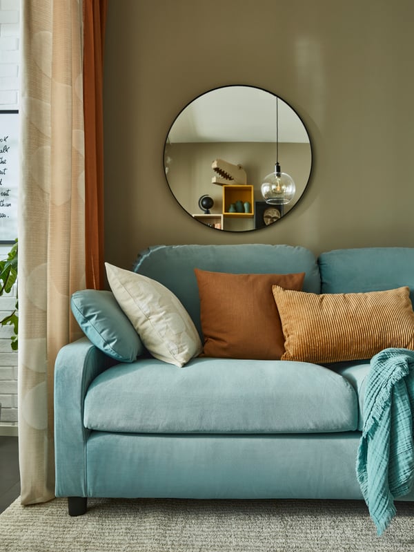 A turquoise sofa with brown and beige cushions, a beige rug, beige and orange curtains, a large round mirror.