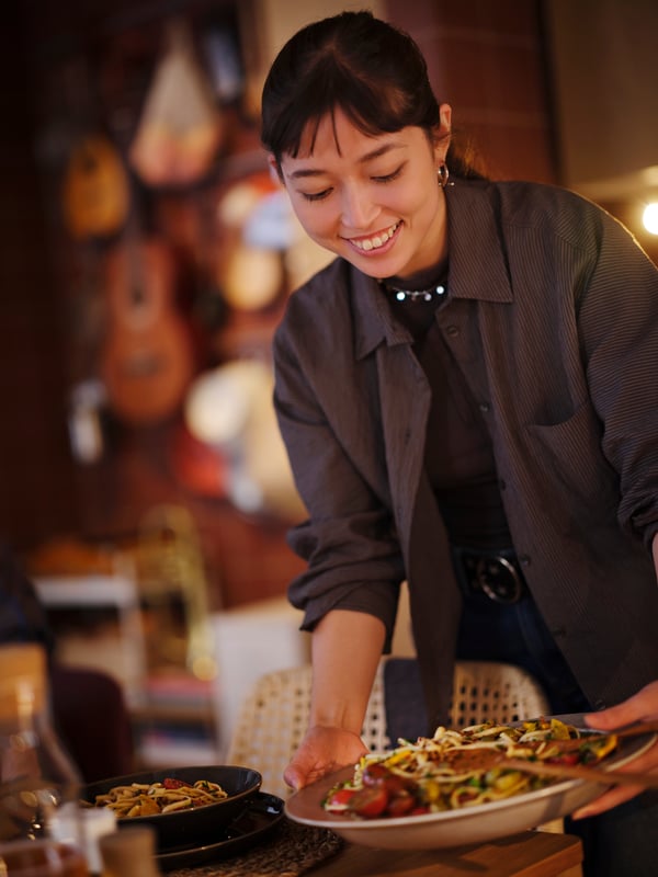 A smiling person in a brown shirt is holding a beige STOCKHOLM 2025 serving bowl with food, about to place it on a table.
