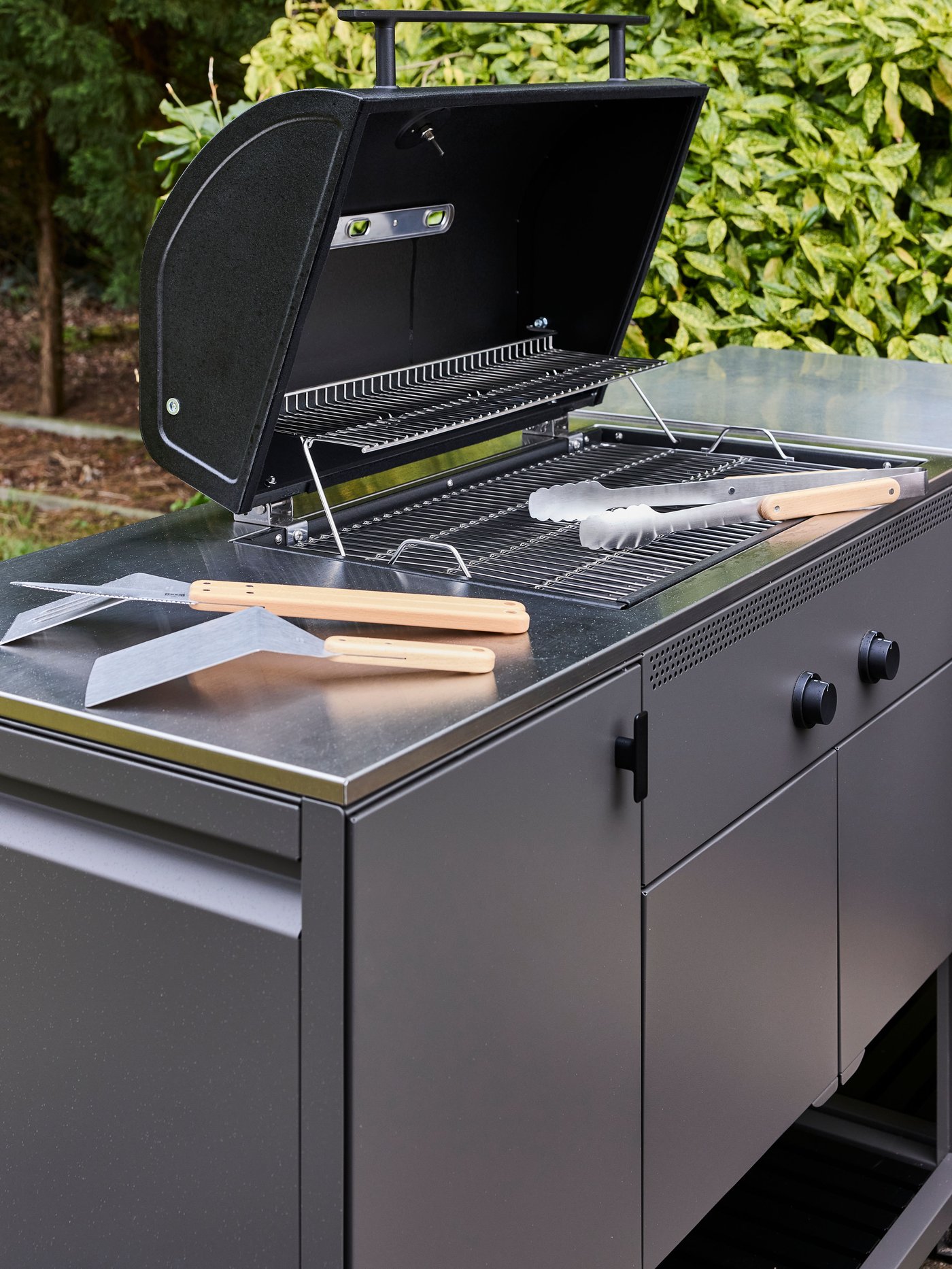 Gasbarbecue met open klep op buitenmeubel, met grillrooster en BBQ-tools, omringd door groen in de tuin.