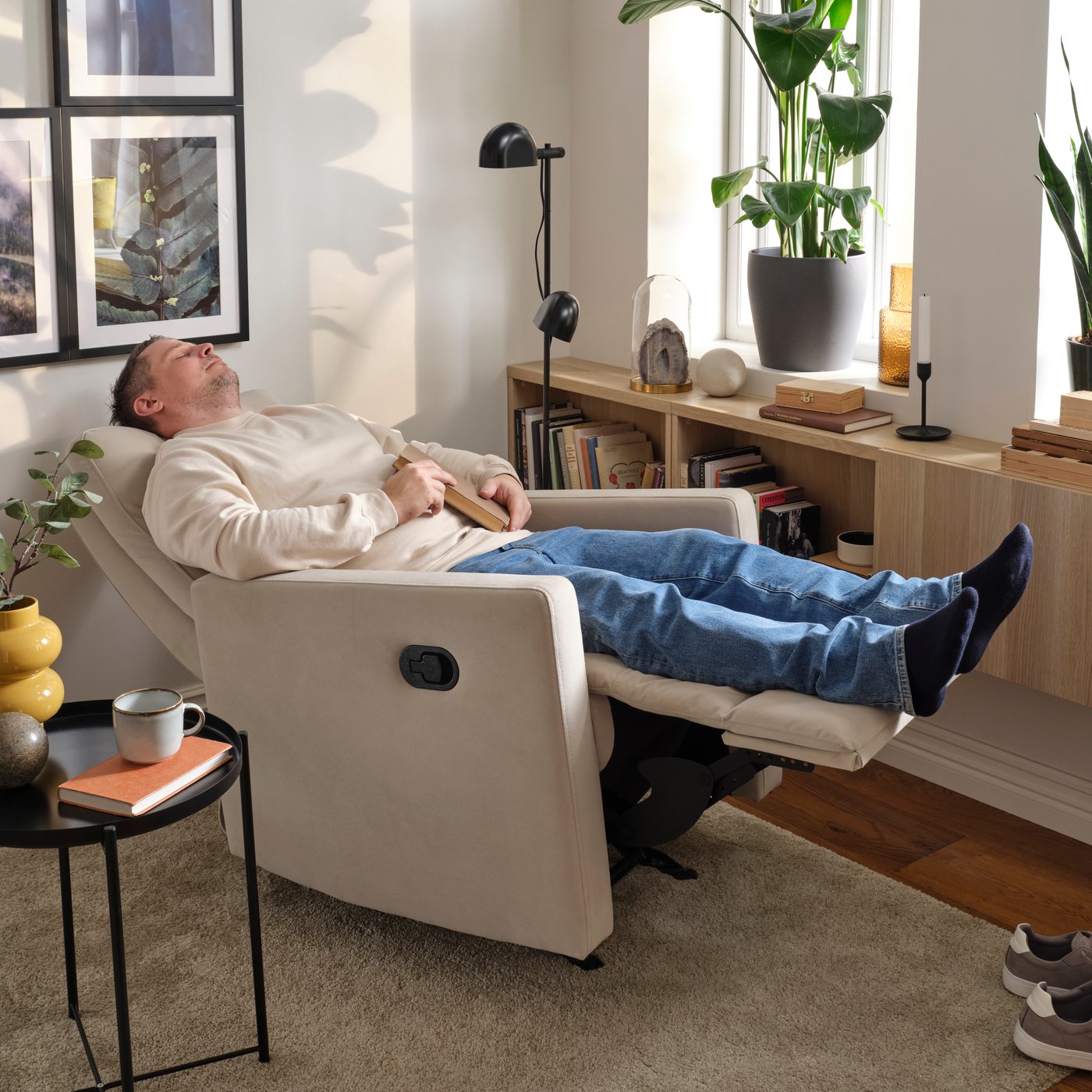 Person relaxing in a recliner with legs raised in a cozy living room decorated with plants, bookshelves, and warm lighting.