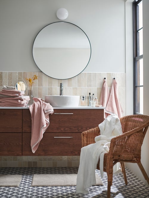 A white bathroom with an AGEN rattan chair beside a wash basin unit that has pink towels on it and a LINDBYN mirror above.