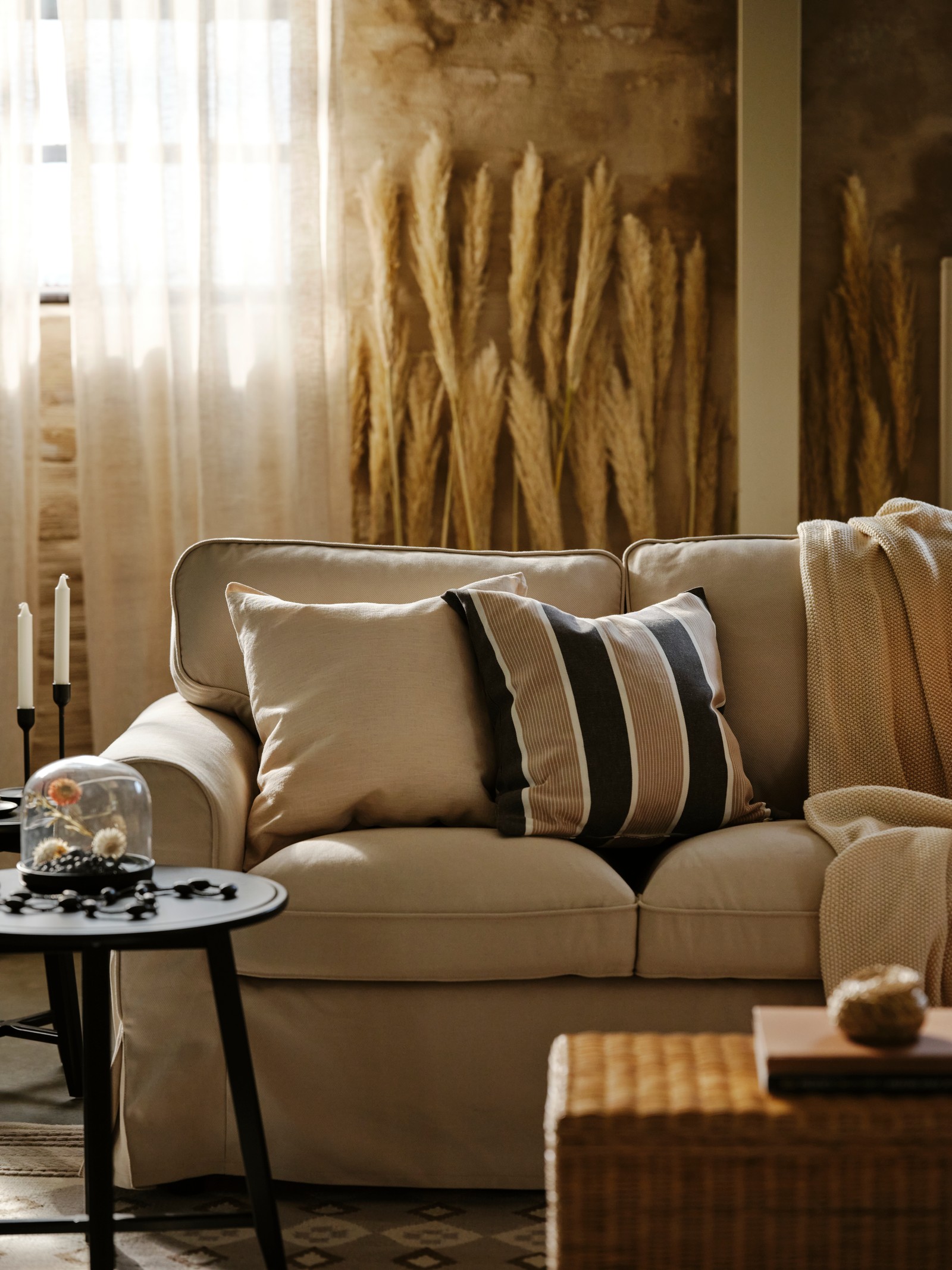 Krem sofa s prugastim i jednobojnim jastucima i tankim ćebetom, crni pomoćni stočići su u blizini i beli prozor sa zavesama je iza.