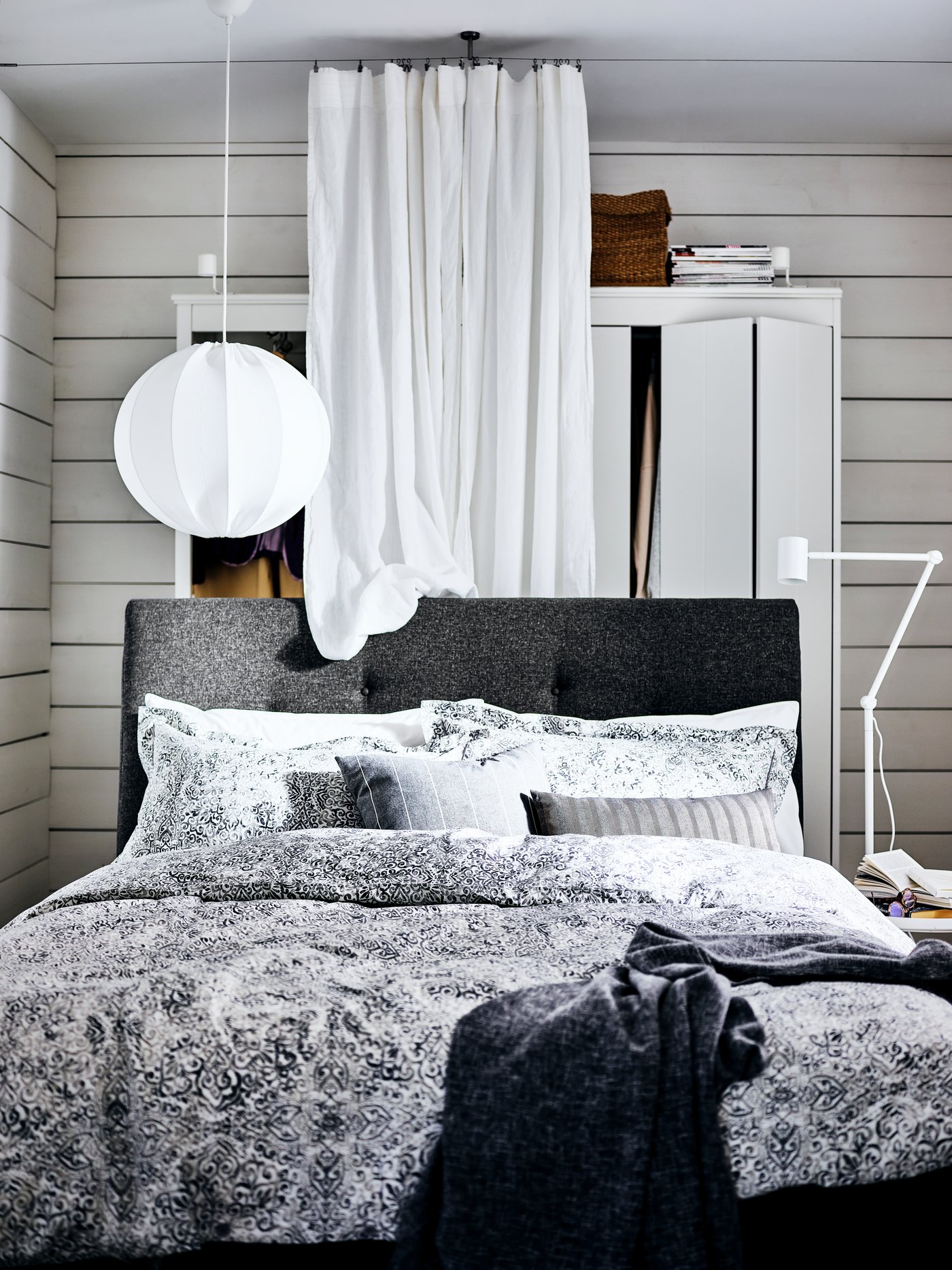 A dark grey IDANÄS upholstered bed with a wardrobe and a white curtain behind it and a NYMÅNE reading lamp beside it.