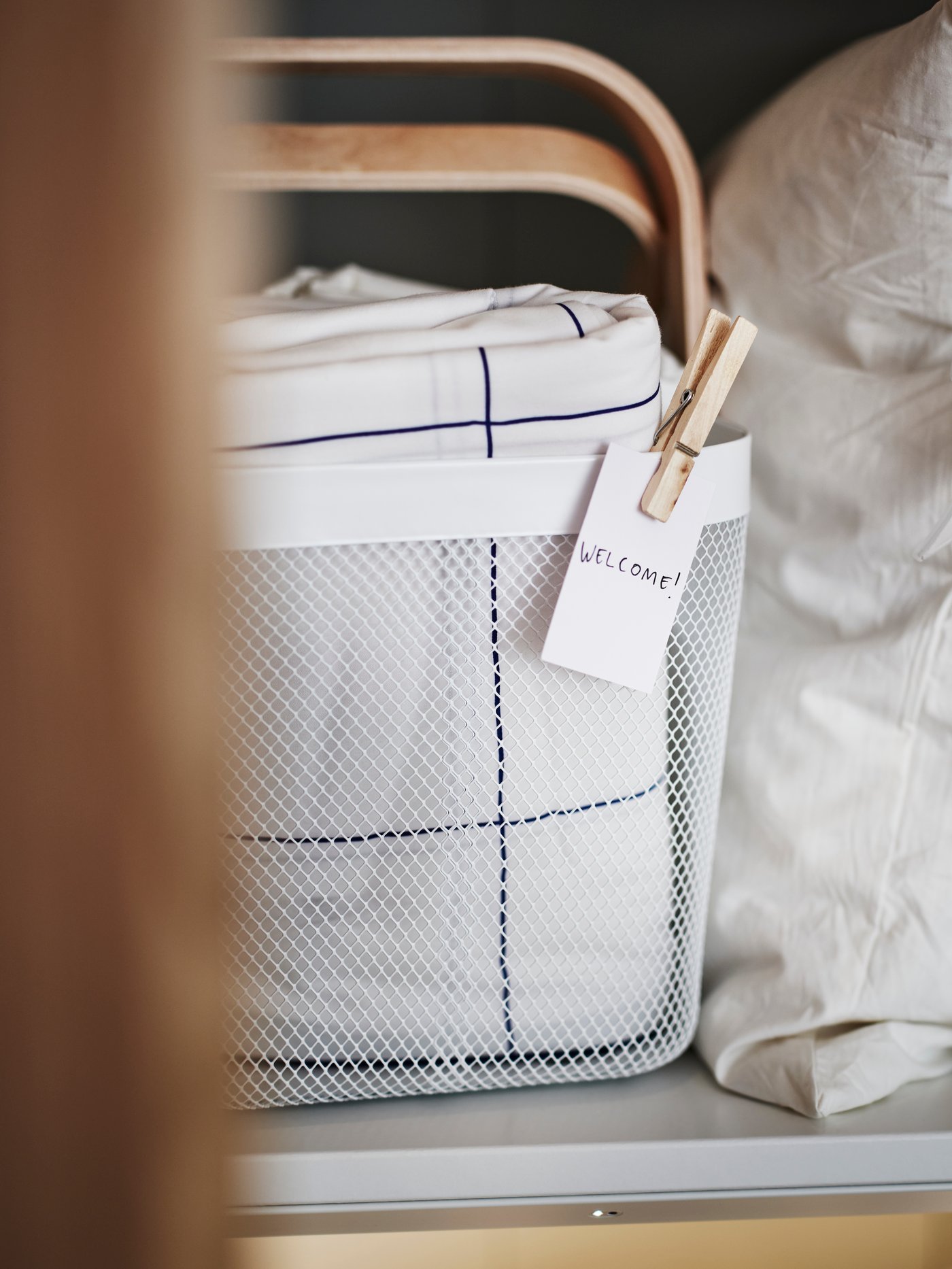 A white RISATORP basket with a label on the front, on a shelf holding folded white and blue check BREDVECKLARE bed linen.