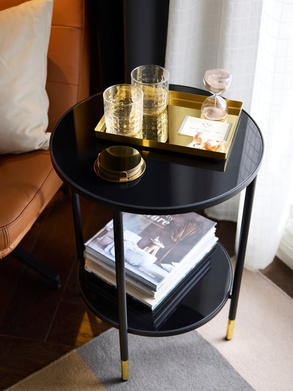 A brass-coloured STOCKHOLM 2025 rectangular tray holds two glasses, wrapped candy and a welcome note on a black side table.