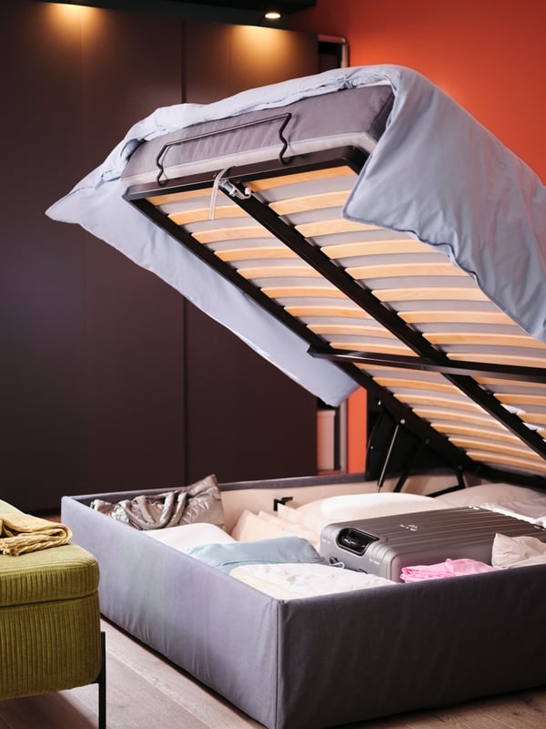 An BLÅFJÄLLET ottoman divan bed with the top open showing the storage area which holds a suitcase, bedding and other items.
