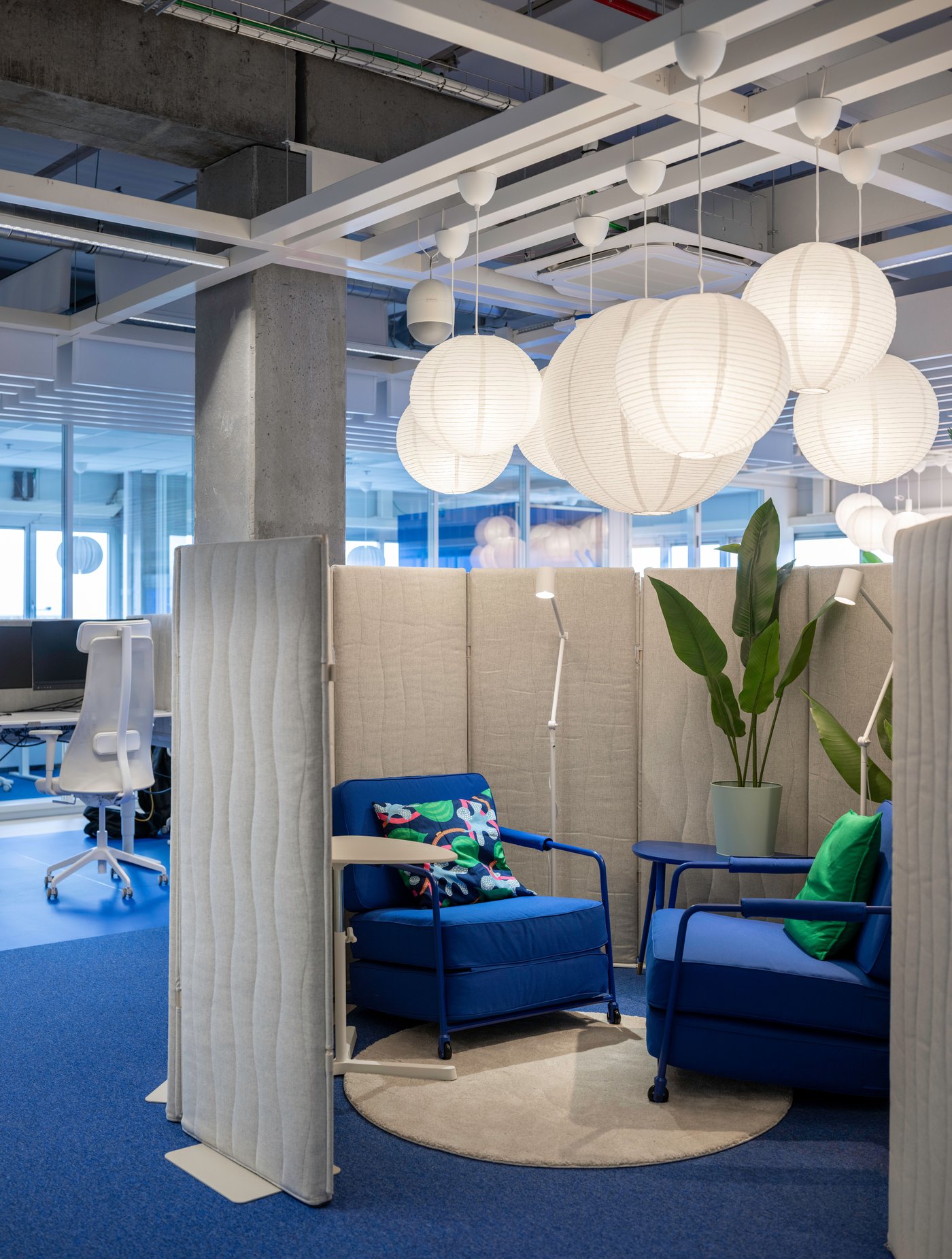 Des fauteuils bleus dans un coin salon de bureau cosy, avec des séparations acoustiques et des suspensions sphériques blanches.