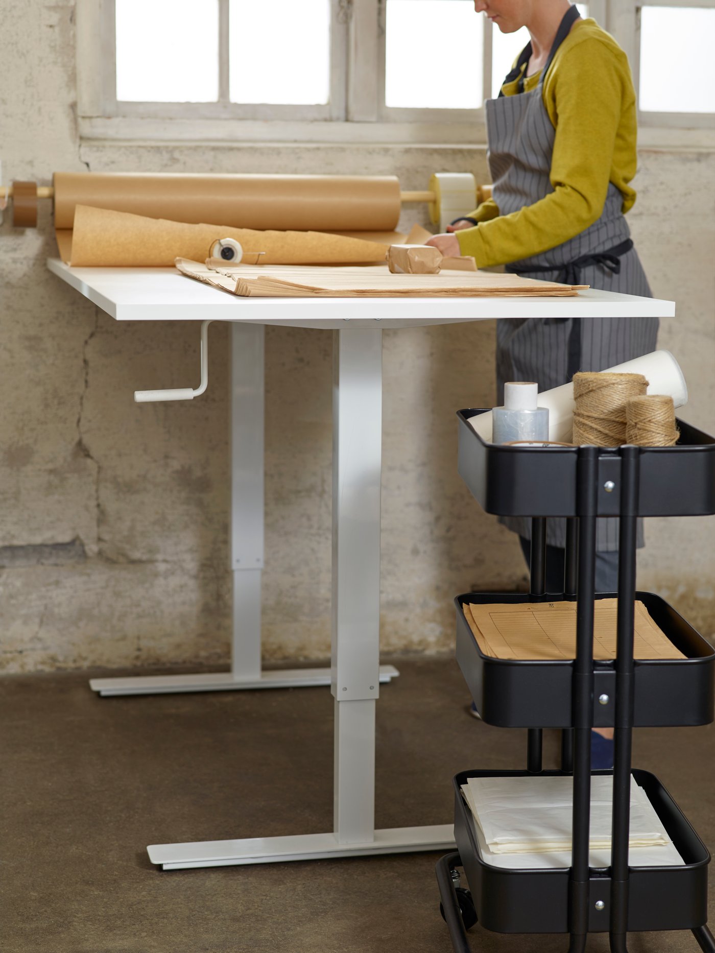 A white RÅSKOG trolley with three levels holding pens, paper and other items, beside a children’s desk and a swivel chair.