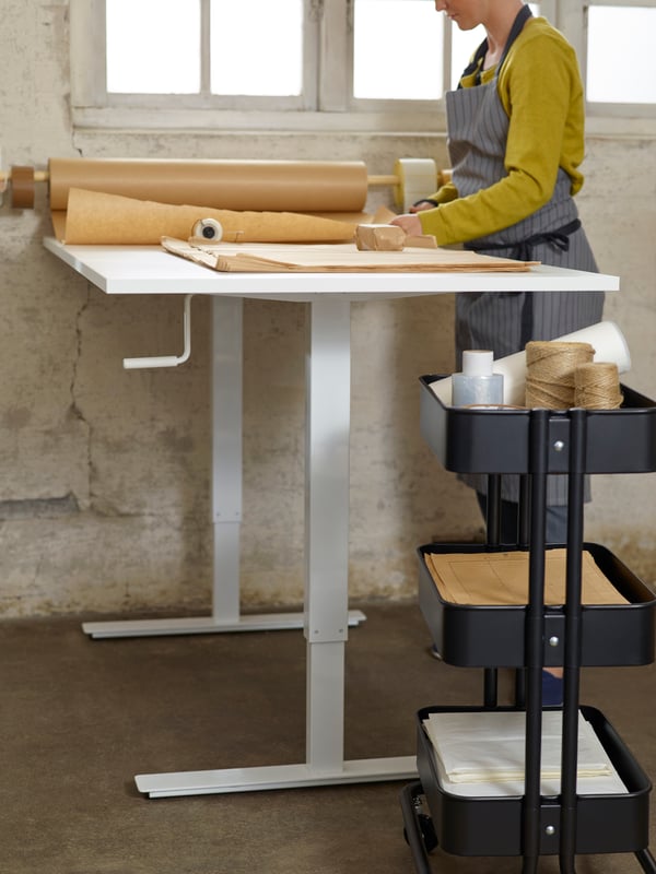 A white RÅSKOG trolley with three levels holding pens, paper and other items, beside a children’s desk and a swivel chair.