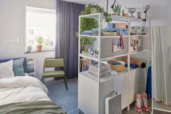 Chambre d’étudiant très éclairée. Un rangement au milieu de la pièce avec divers articles posés sur les étagères. Une chaise verte à côté d’un lit.