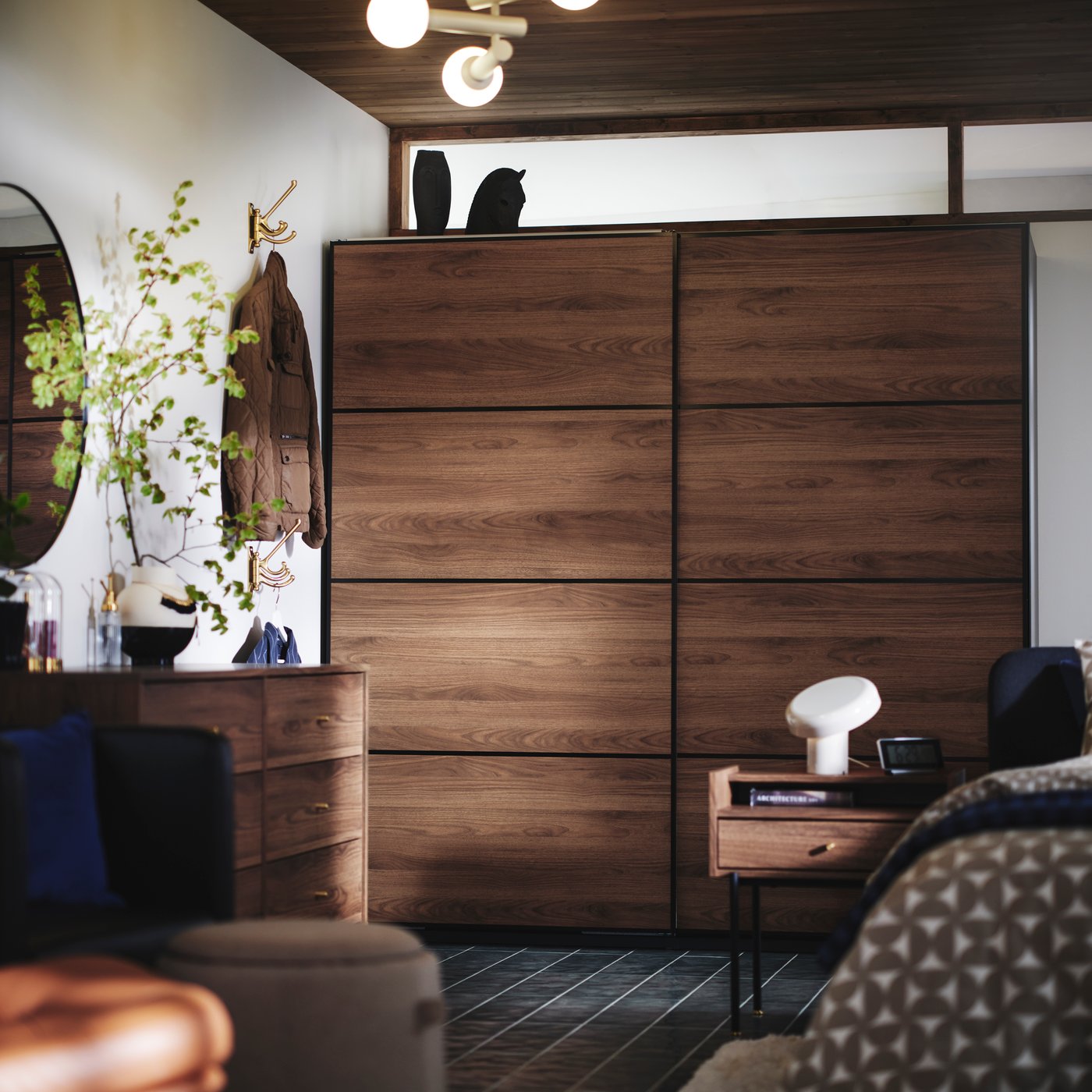The side of a bed, RÅDMANSÖ bedside table and PAX / MEHAMN wardrobe in dark wood and sliding doors in the background.