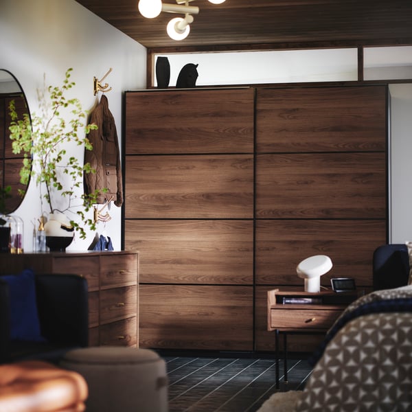The side of a bed, RÅDMANSÖ bedside table and PAX MEHAMN wardrobe in dark wood and sliding doors in the background.