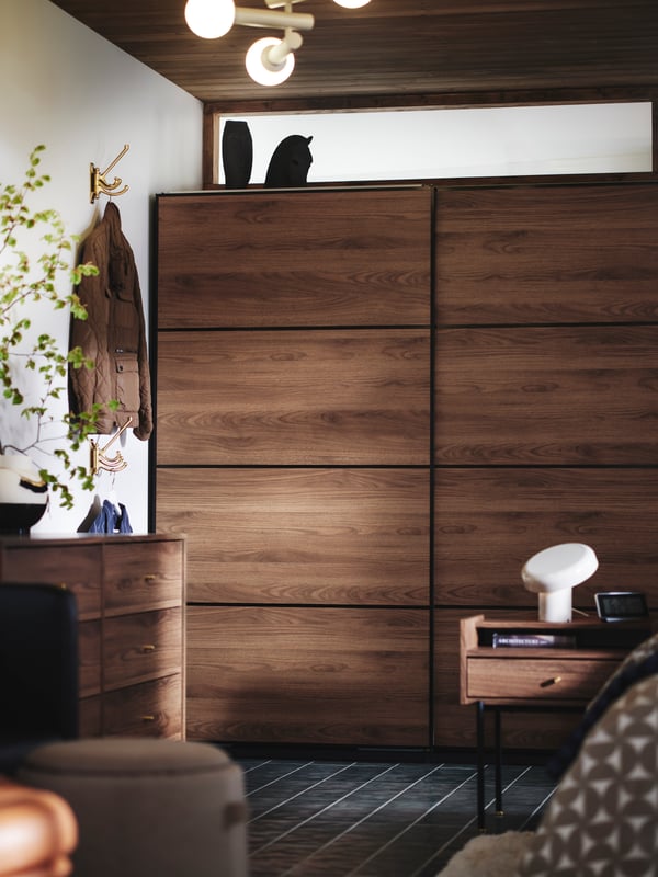 Op de achtergrond een bedrand, het RÅDMANSÖ nachtkastje en de PAX MEHAMN kledingkast in donker hout, met schuifdeuren.