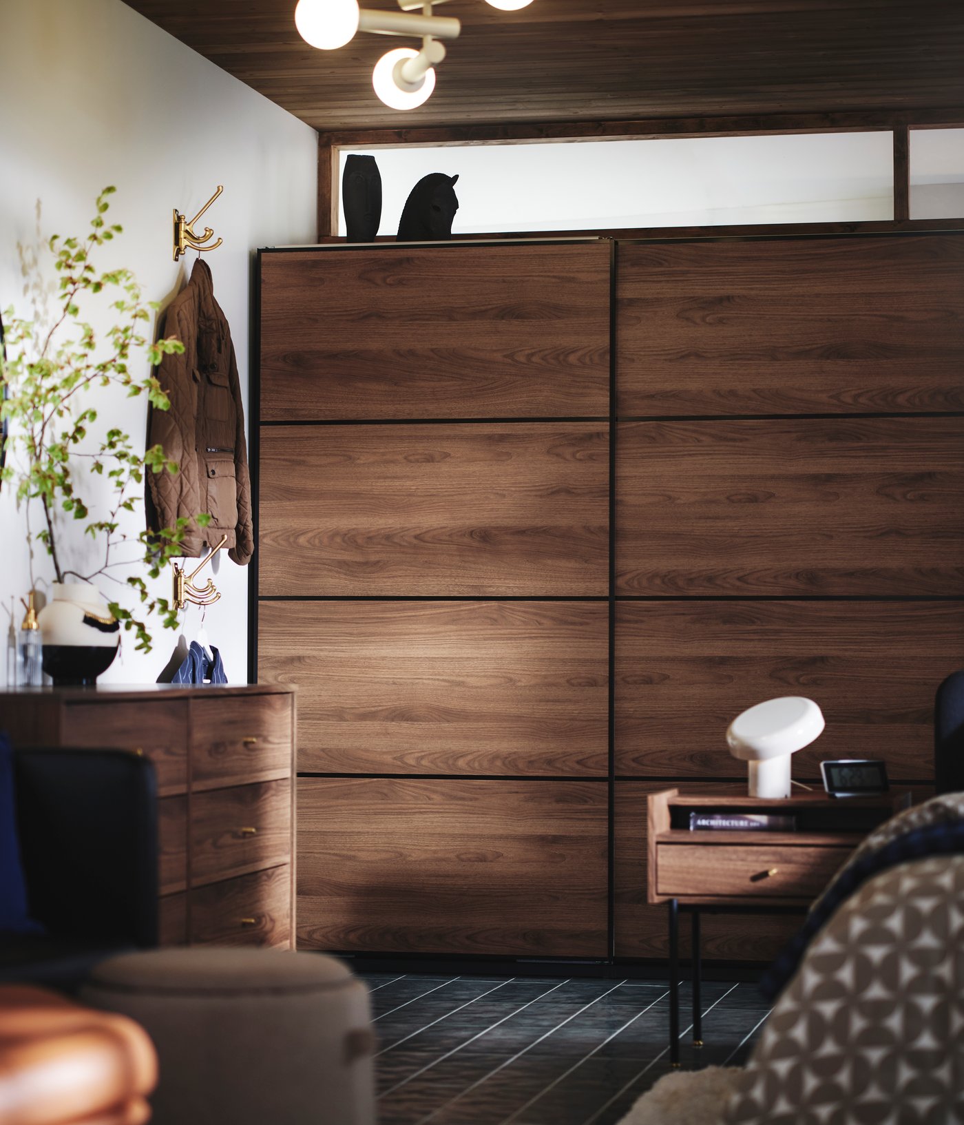 The side of a bed, RÅDMANSÖ bedside table and PAX / MEHAMN wardrobe in dark wood and sliding doors in the background.