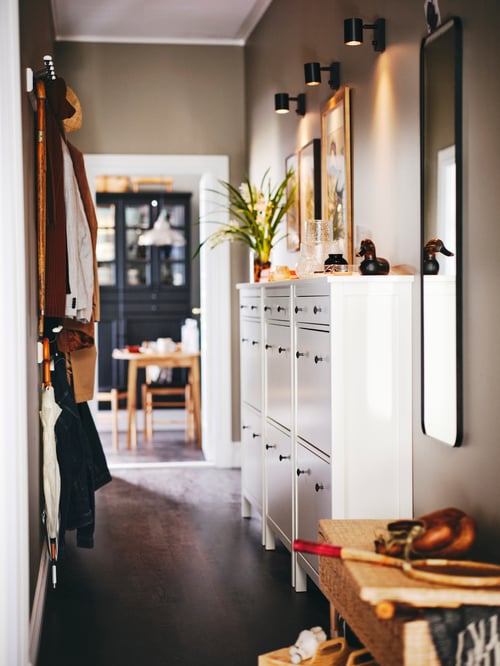 Flurbereich mit mehreren HEMNES Schuhschränken mit zwei Fächern, einem LINDBYN Spiegel und LANDKRABBA Hakenleisten, an denen ein Schirm und Kleidungsstücke zu sehen sind.