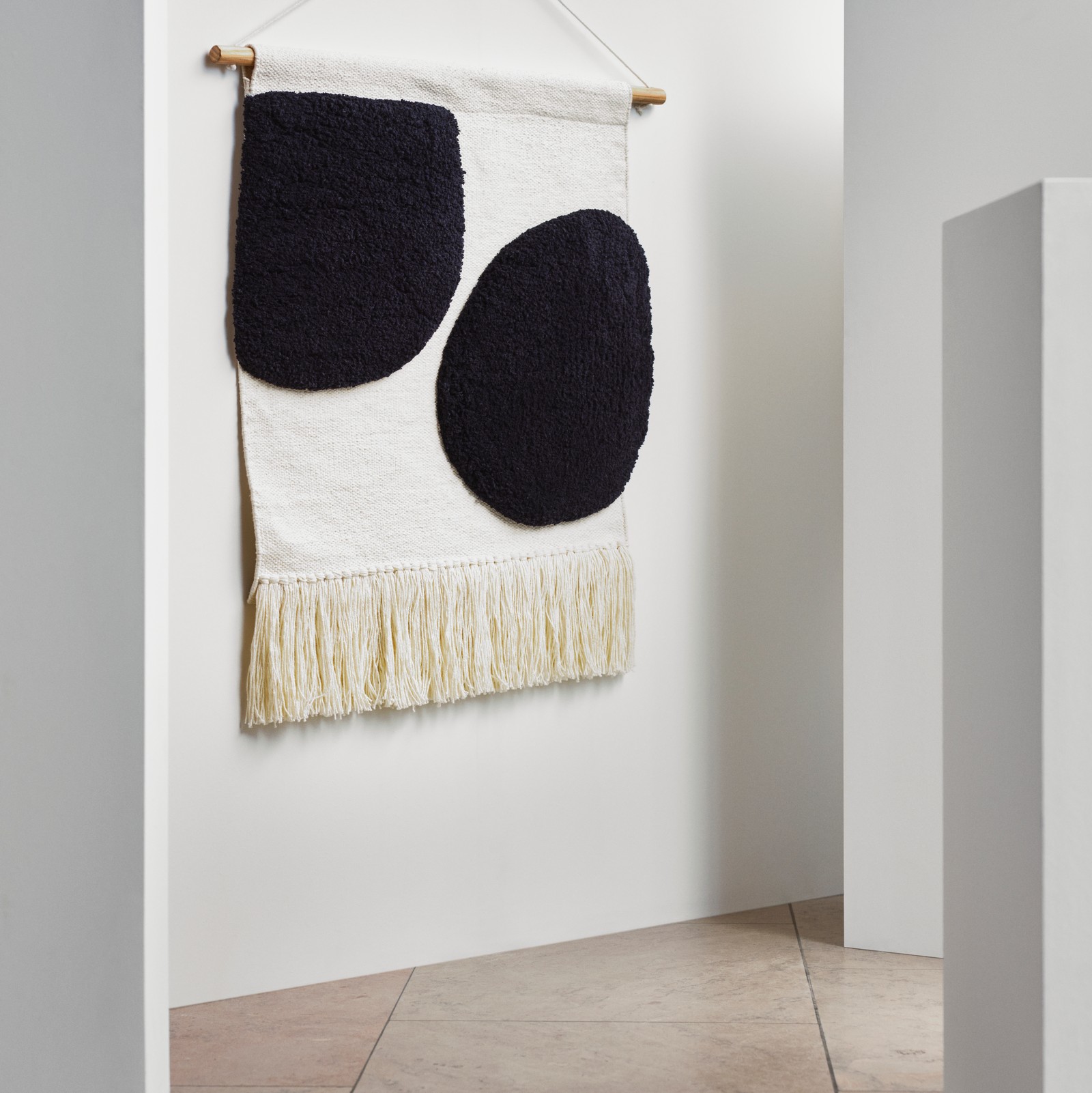 A BLANDSKOG wall tapestry features two black circles on a beige background, and hangs from a plain white wall.