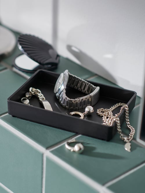 A black SKOGSVIKEN tray stands on a green-tiled bathroom ledge and is filled with various pieces of silver jewellery.