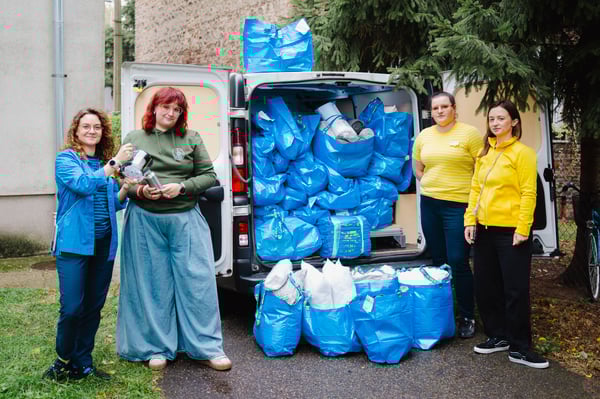 Pracownicy IKEA oraz jedna osoba z fundacji stojąca na tle samochodu dostawczego z otwartym bagażnikiem wypakowanym niebieskiemi torbami z IKEA