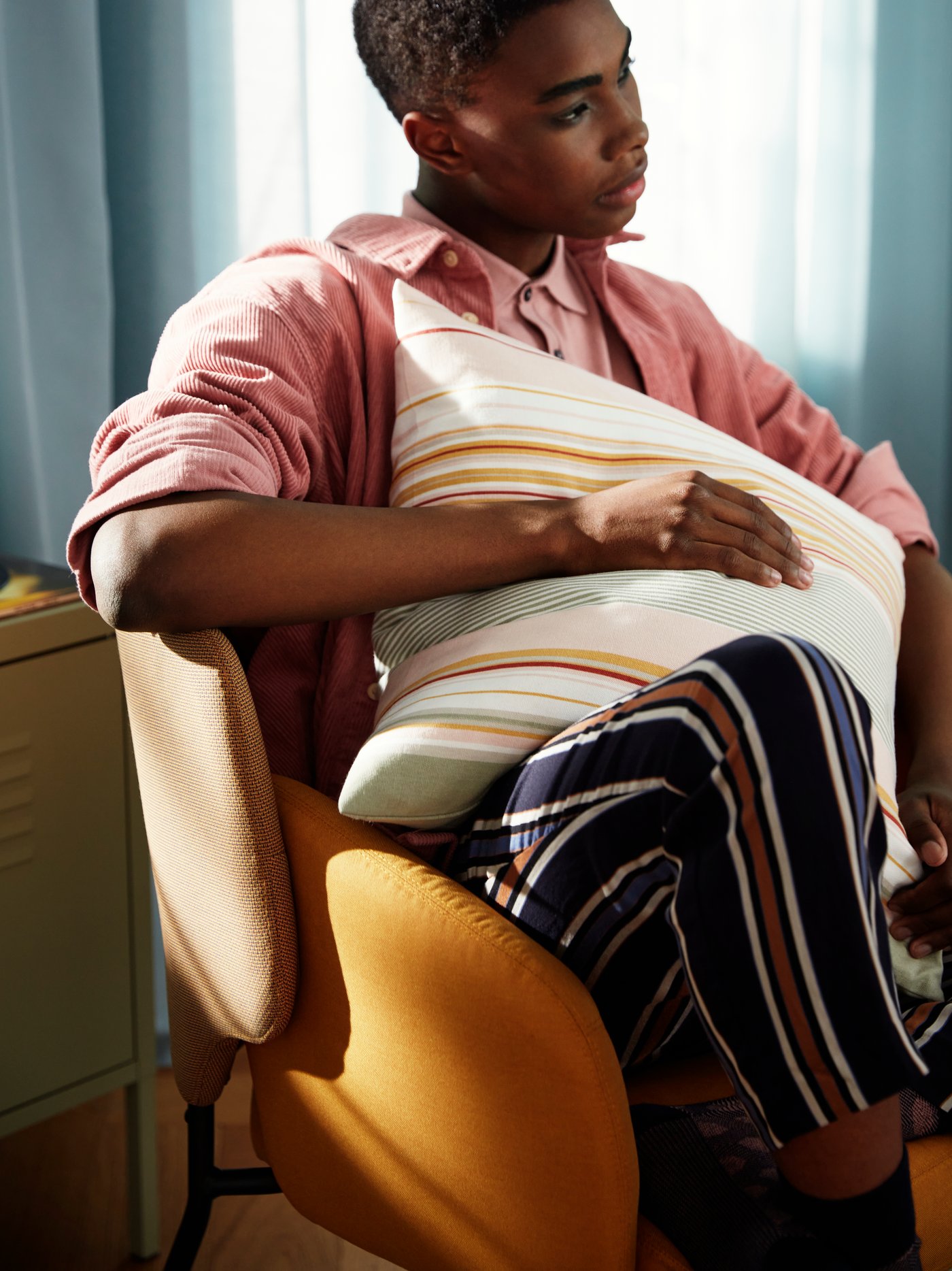 A man sitting in a yellow chair holding a SOLMOTT cushion cover striped in yellow, orange, green and white.
