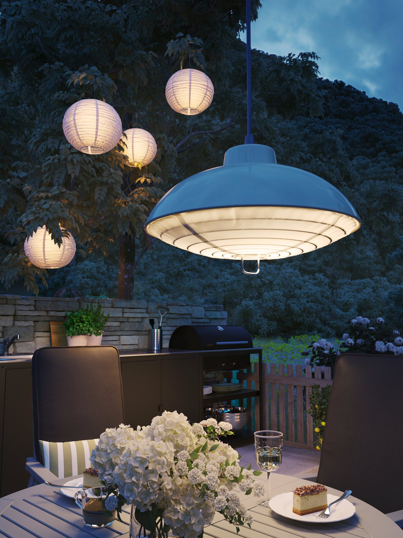 Four round paper lanterns hang on trees while a solar-powered lamp hang above a table with dessert and flowers on it.