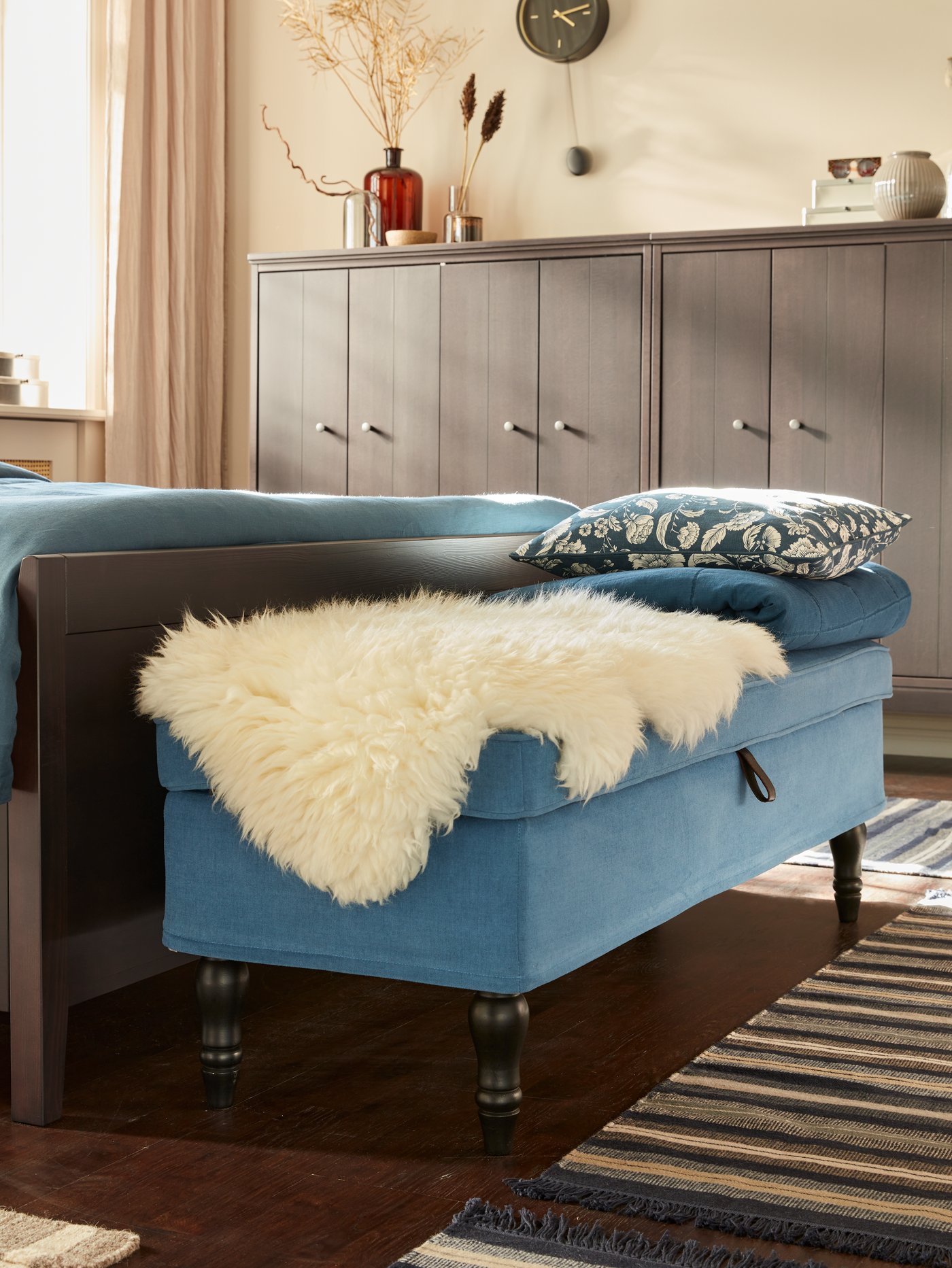 A blue upholstered bench at the foot of a wooden bed, with a white sheepskin rug, blue cushions and throw on it.