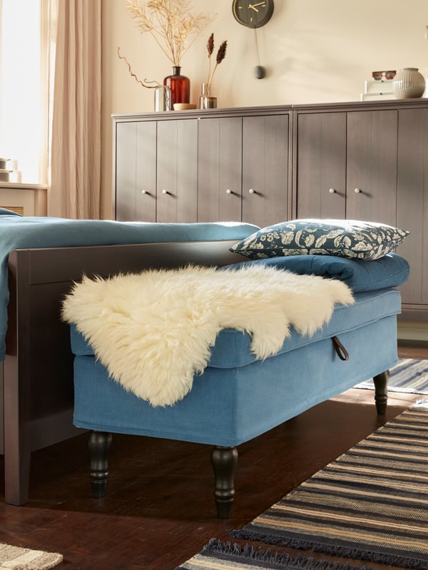 A blue upholstered bench at the foot of a wooden bed, with a white sheepskin rug, blue cushions and throw on it.