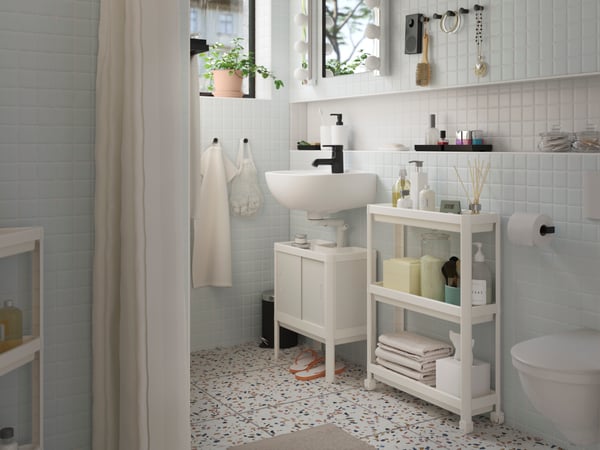 A white LILLTJÄRN/SKATSJÖN bathroom furniture set with a SALJEN tap beside a white VESKEN rolling cart holding toiletries.
