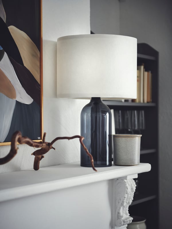 A smoked-glass-and-white TONVIS table lamp placed next to framed art on a mantlepiece in a light room.