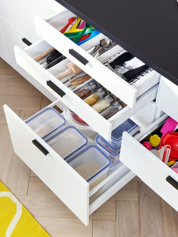 Une cuisine avec des tiroirs blancs ouverts sur des boîtes de conservation pour aliments en plastique et des ustensiles de cuisine.