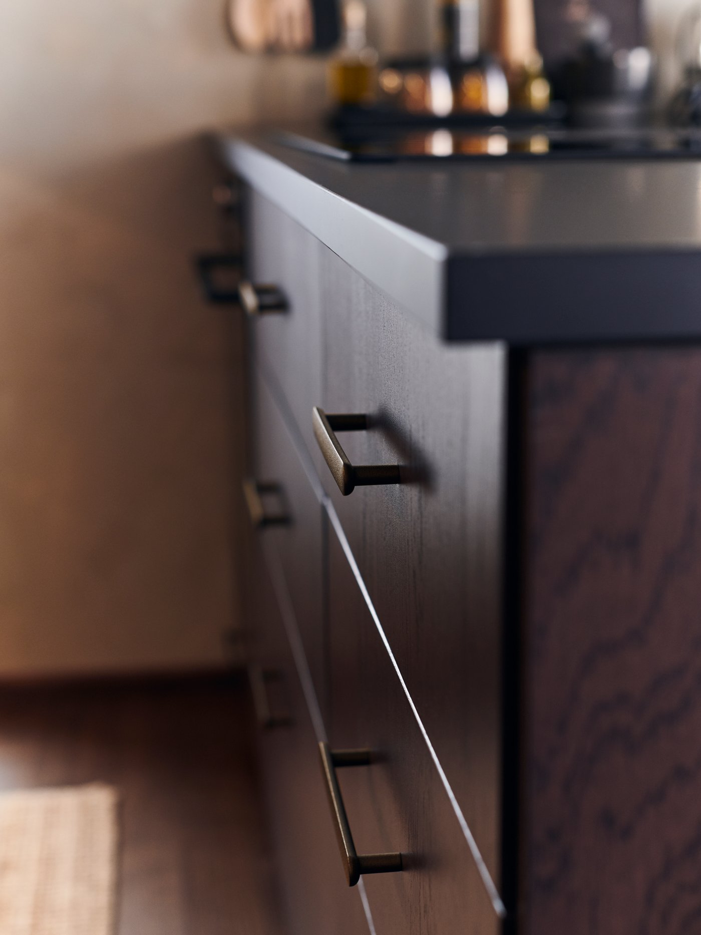 وحدة مطبخ بتكسية خشب غامق تضم أدراجاً ومقابض باللون البرونزي. سطح عمل EKBACKEN وموقد مدمج.