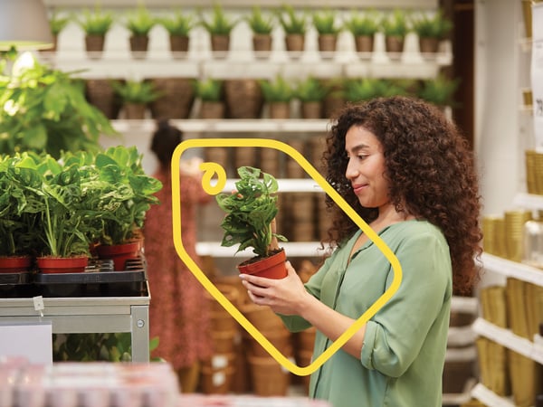 Woman holding a green potted plant in a terracotta pot, looking at the plant section in IKEA with shelves full of plants in the background.