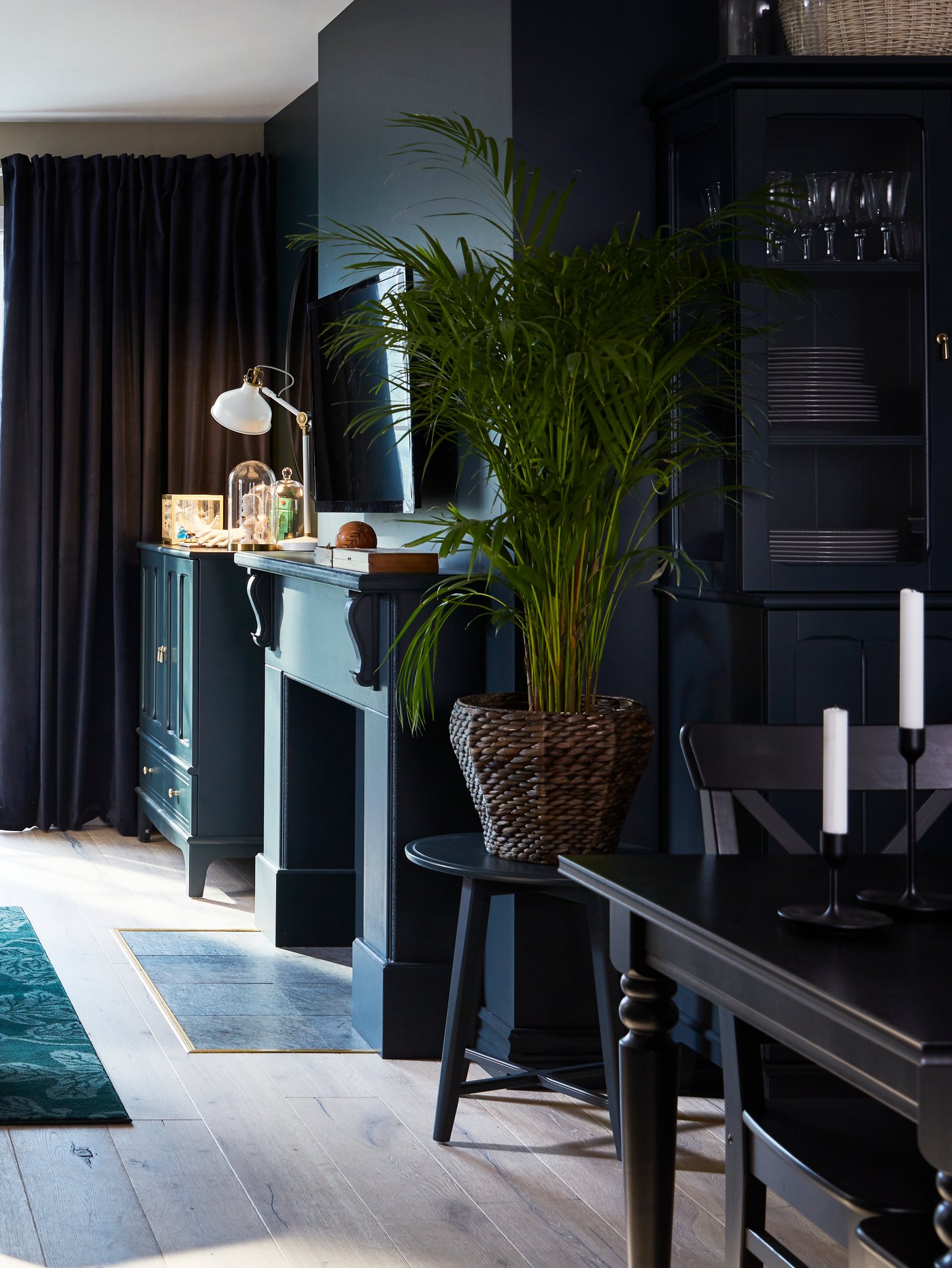 Salón oscuro con cortinas opacas grises, mesa y sillas negras, chimenea, mueble para TV y maceta.