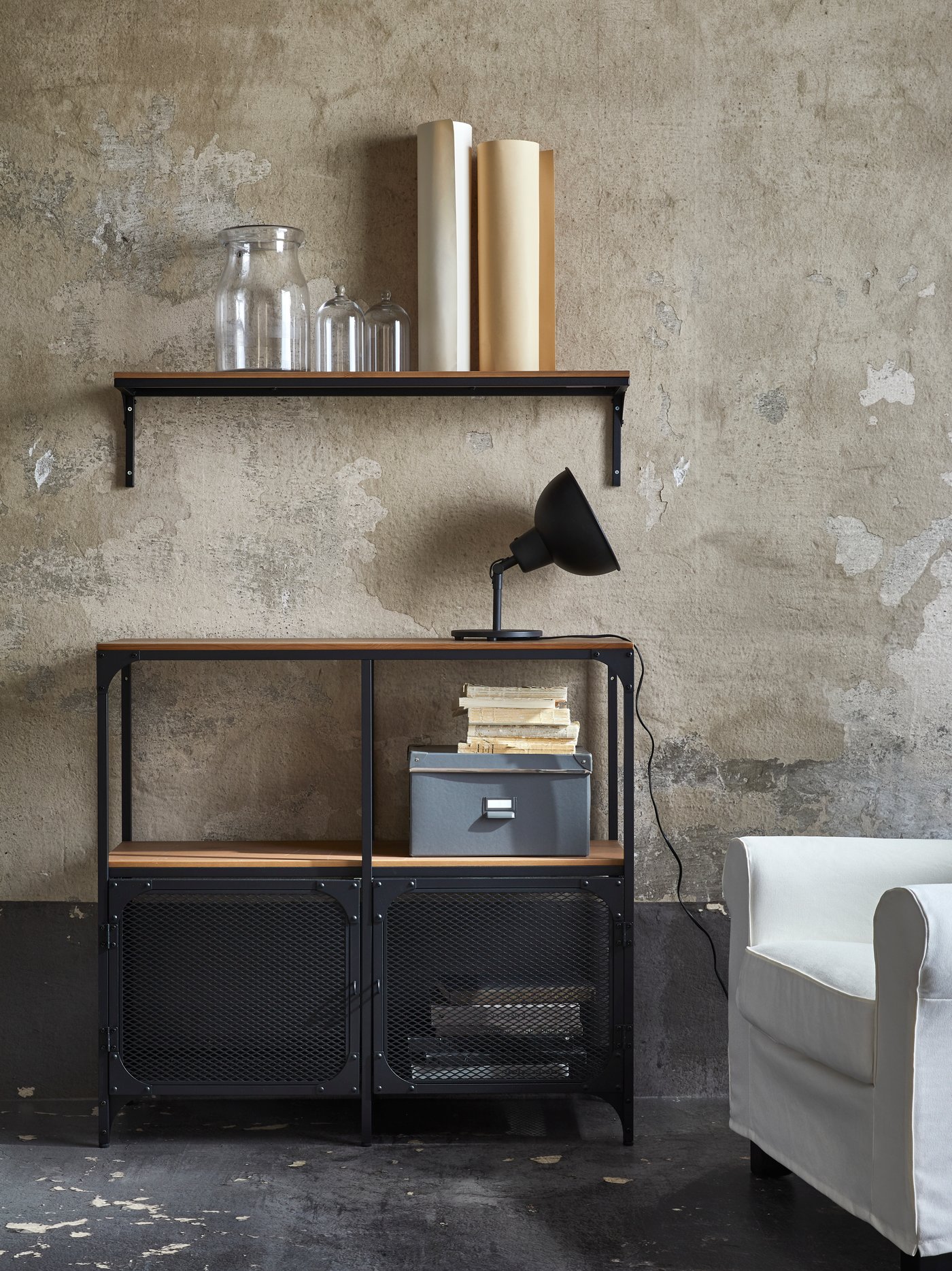 Salón con estantería de metal y madera, estante de pared, cajón, jarrón de vidrio, lámpara de pared y sillón blanco. 