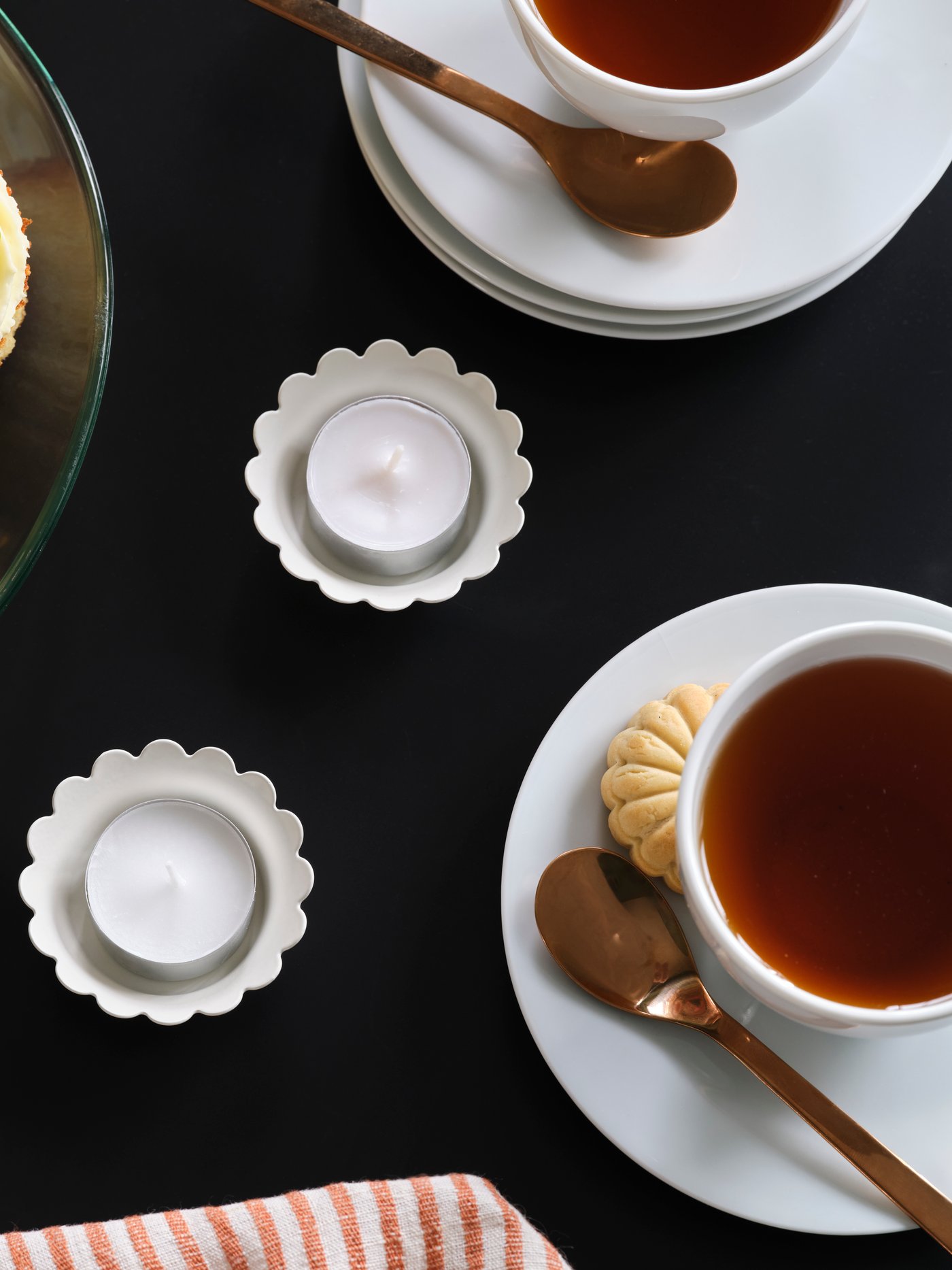 Two SNÖSISKA candle holders in off-white stand on a black table surrounded by two cups of tea and a cake stand with cookies.